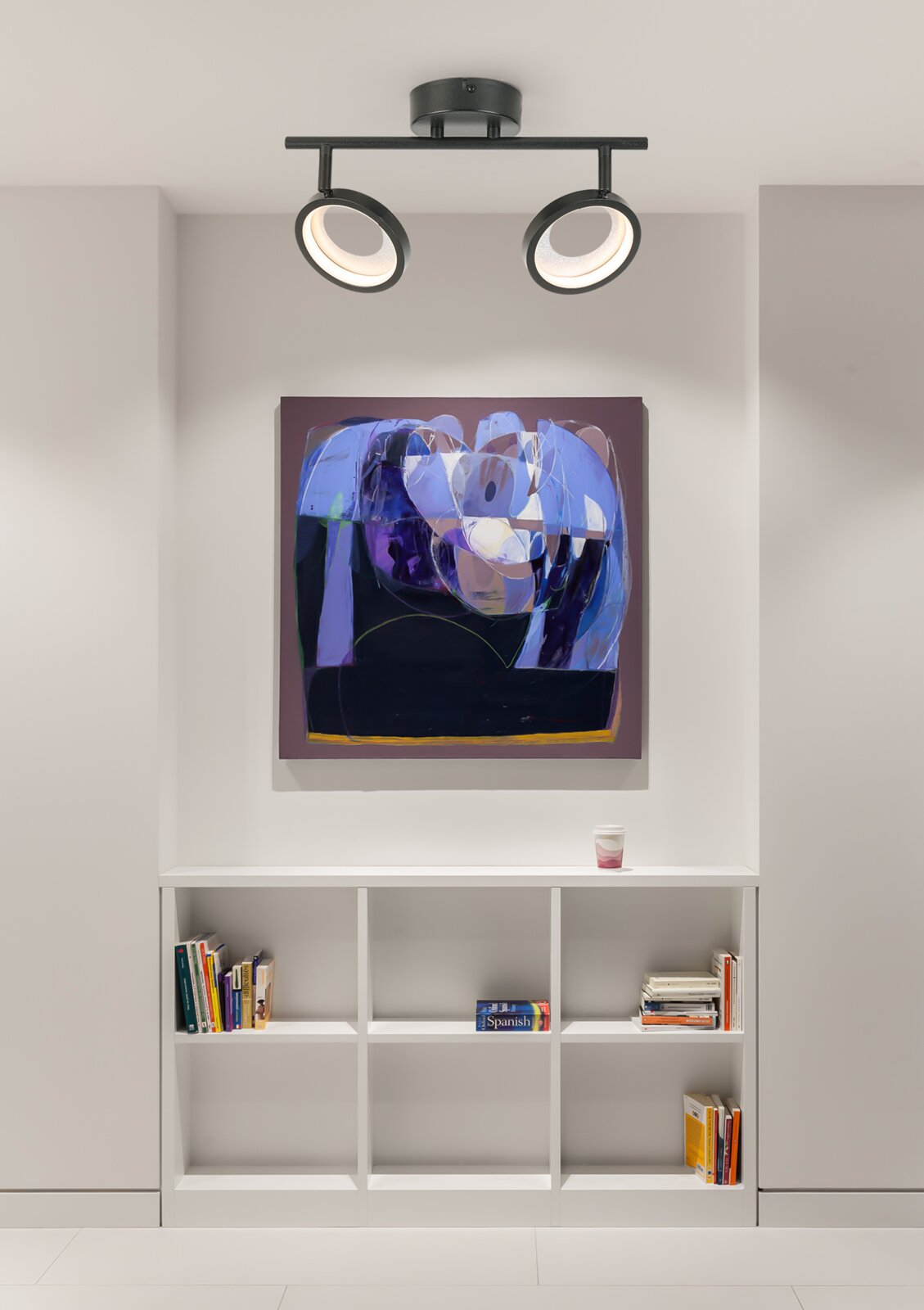 Moderne led-spots aan het plafond, gezien vanaf de voorkant, verlichten een abstract schilderij boven een witte boekenkast.