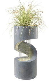 Moderner grauer Gartenbrunnen in Betonoptik mit S-förmiger Wasserlauf-Rinne und Pflanzschale mit Ziergras, Wasser fließt in ein rundes Becken, Frontansicht leicht seitlich vor weißem Hintergrund.