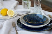Blau glasierte Keramikschale auf weißem Teller und blauem Speiseteller auf gedecktem Tisch mit Zitrone und Wassergläsern, fotografiert aus leicht schräger Seitenperspektive (3/4-Ansicht).