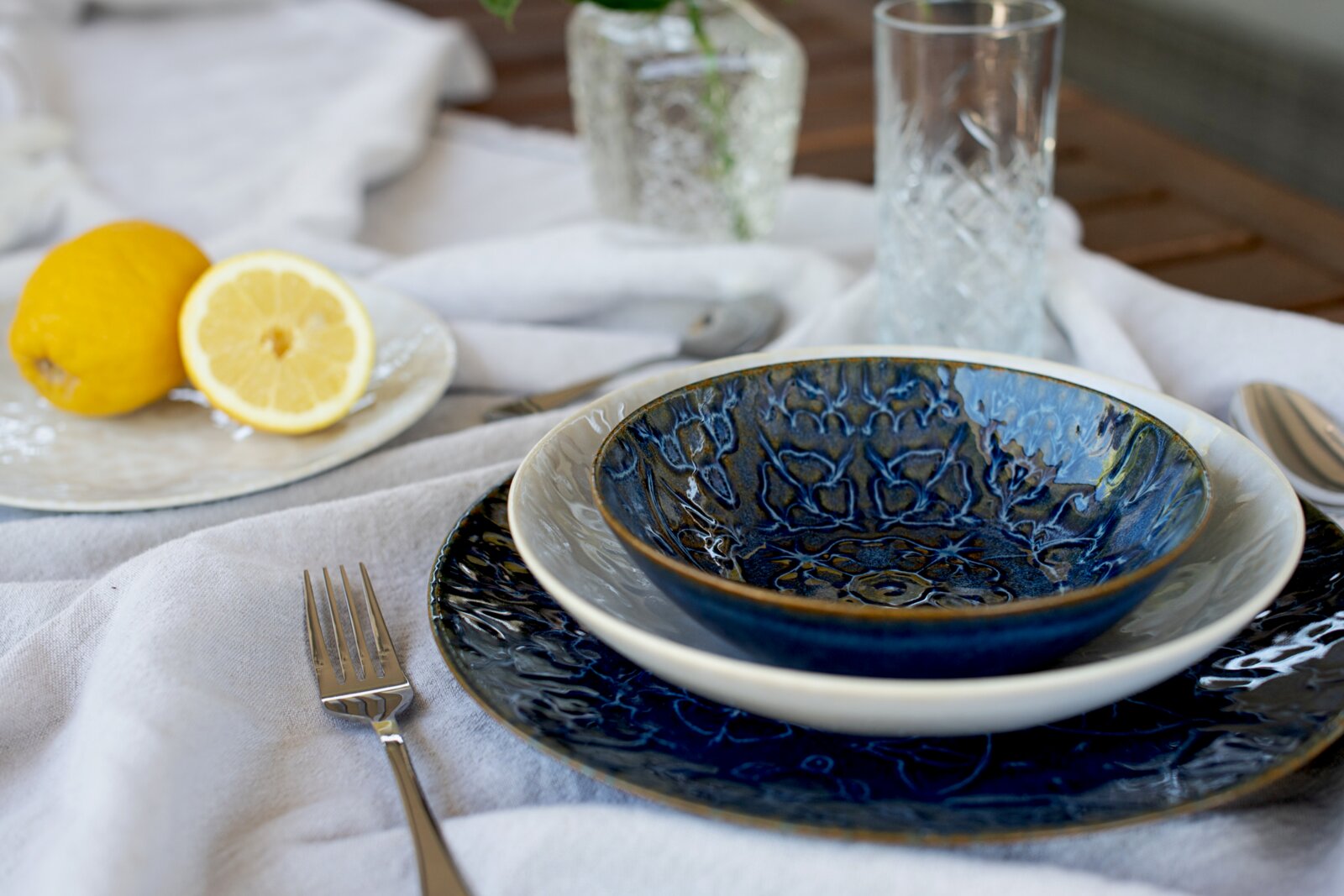 Blau glasierte Keramikschale auf weißem Teller und blauem Speiseteller auf gedecktem Tisch mit Zitrone und Wassergläsern, fotografiert aus leicht schräger Seitenperspektive (3/4-Ansicht).