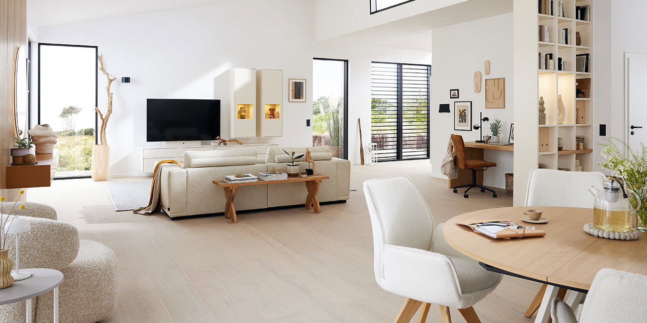 Das Wohnzimmer ist hell und modern eingerichtet. Ein großes Sofa lädt zum Entspannen ein. Es gibt einen Fernseher und viel Platz. Ein Esstisch mit Stühlen steht im Raum, ideal für Mahlzeiten. Helle Möbel und große Fenster lassen den Raum freundlich wirken