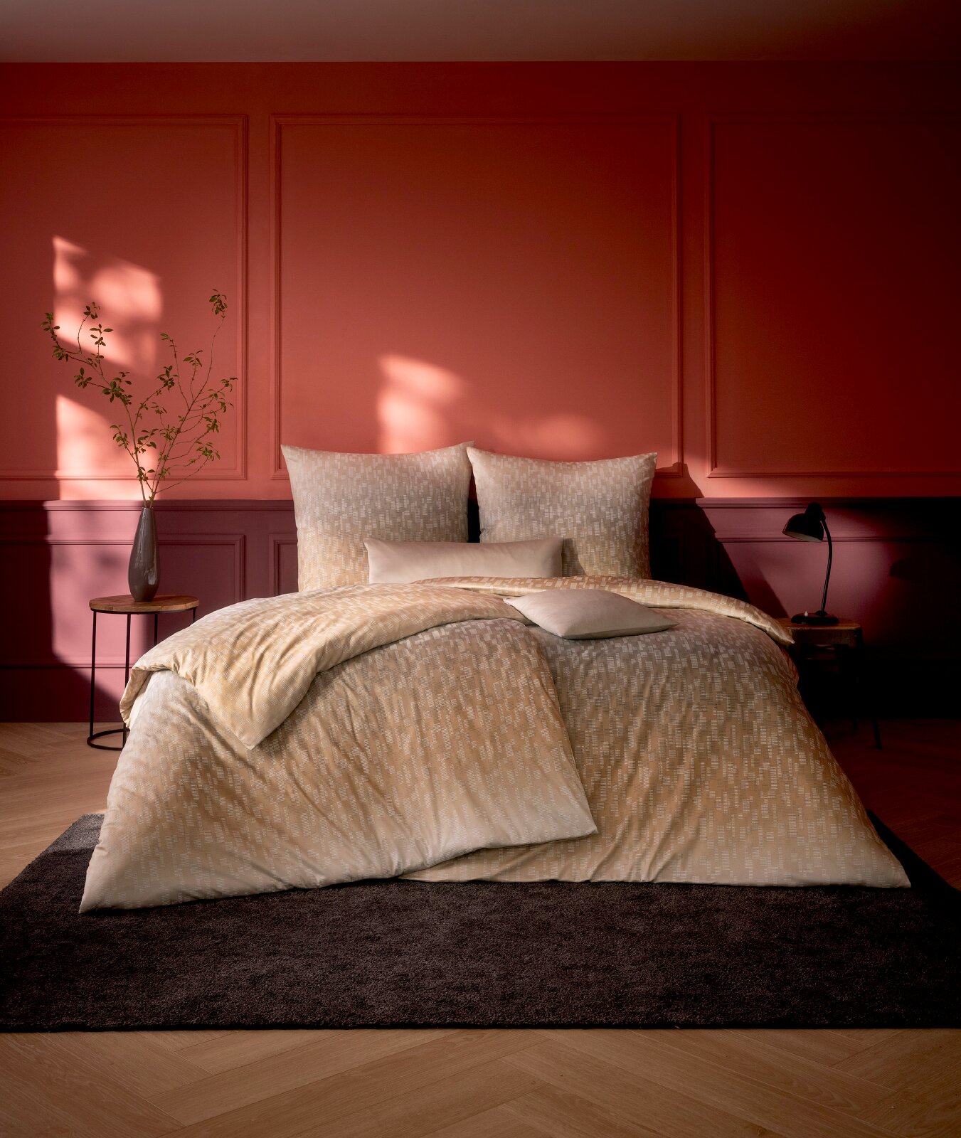 Beige Bettwäsche mit dezentem grafischem Muster auf einem Doppelbett, frontal in stimmungsvoll beleuchtetem Schlafzimmer vor roter Wand präsentiert