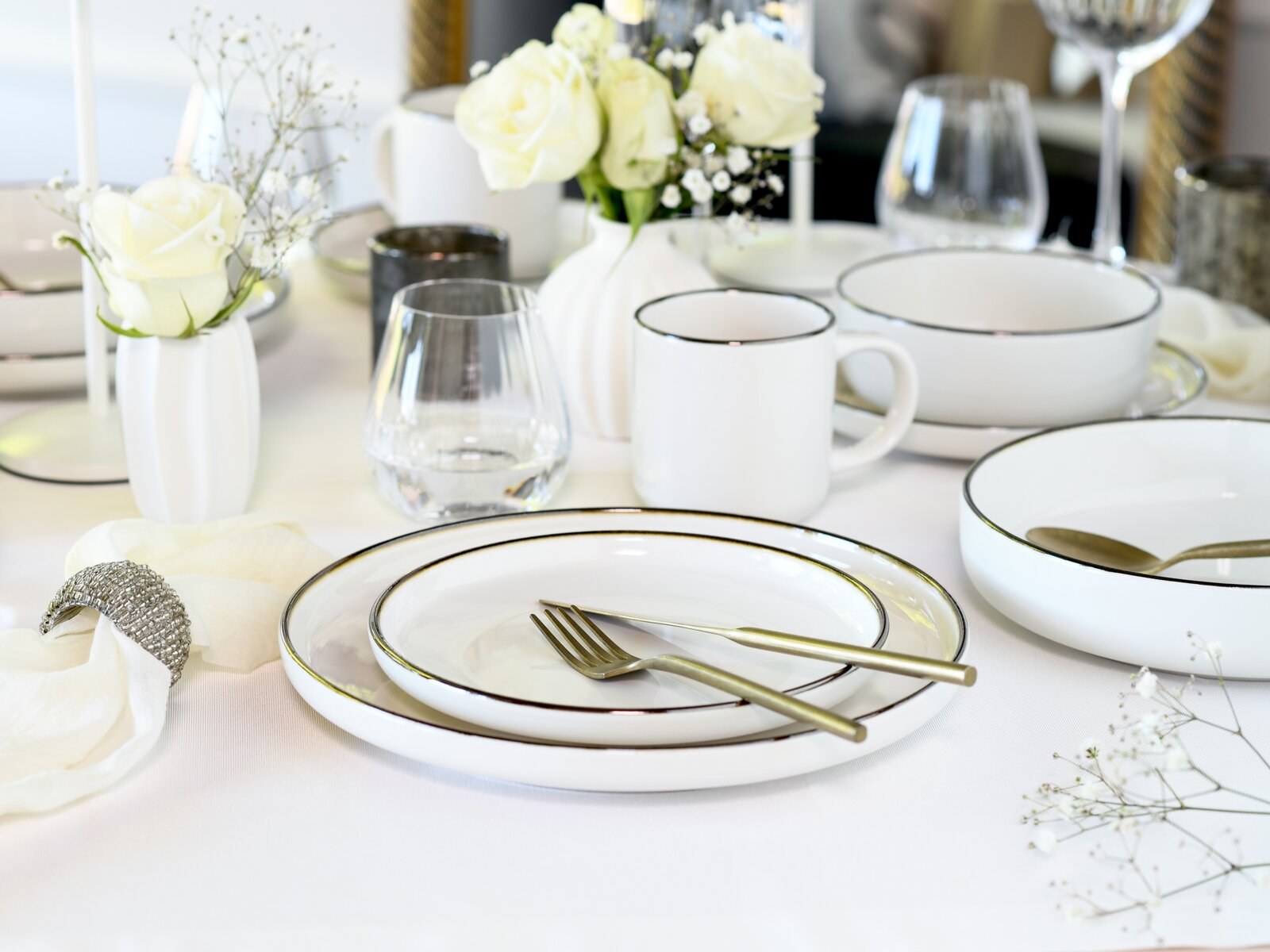 Elegant gedeckter Tisch mit weißem Porzellan-Geschirr (Teller, Schalen, Tasse) mit feinem dunklem Rand, goldfarbenem Besteck, Gläsern und weißen Rosen in Vasen, aufgenommen aus leicht erhöhter schräger Perspektive.