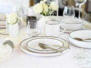 Elegant gedeckter Tisch mit weißem Porzellan-Geschirr (Teller, Schalen, Tasse) mit feinem dunklem Rand, goldfarbenem Besteck, Gläsern und weißen Rosen in Vasen, aufgenommen aus leicht erhöhter schräger Perspektive.