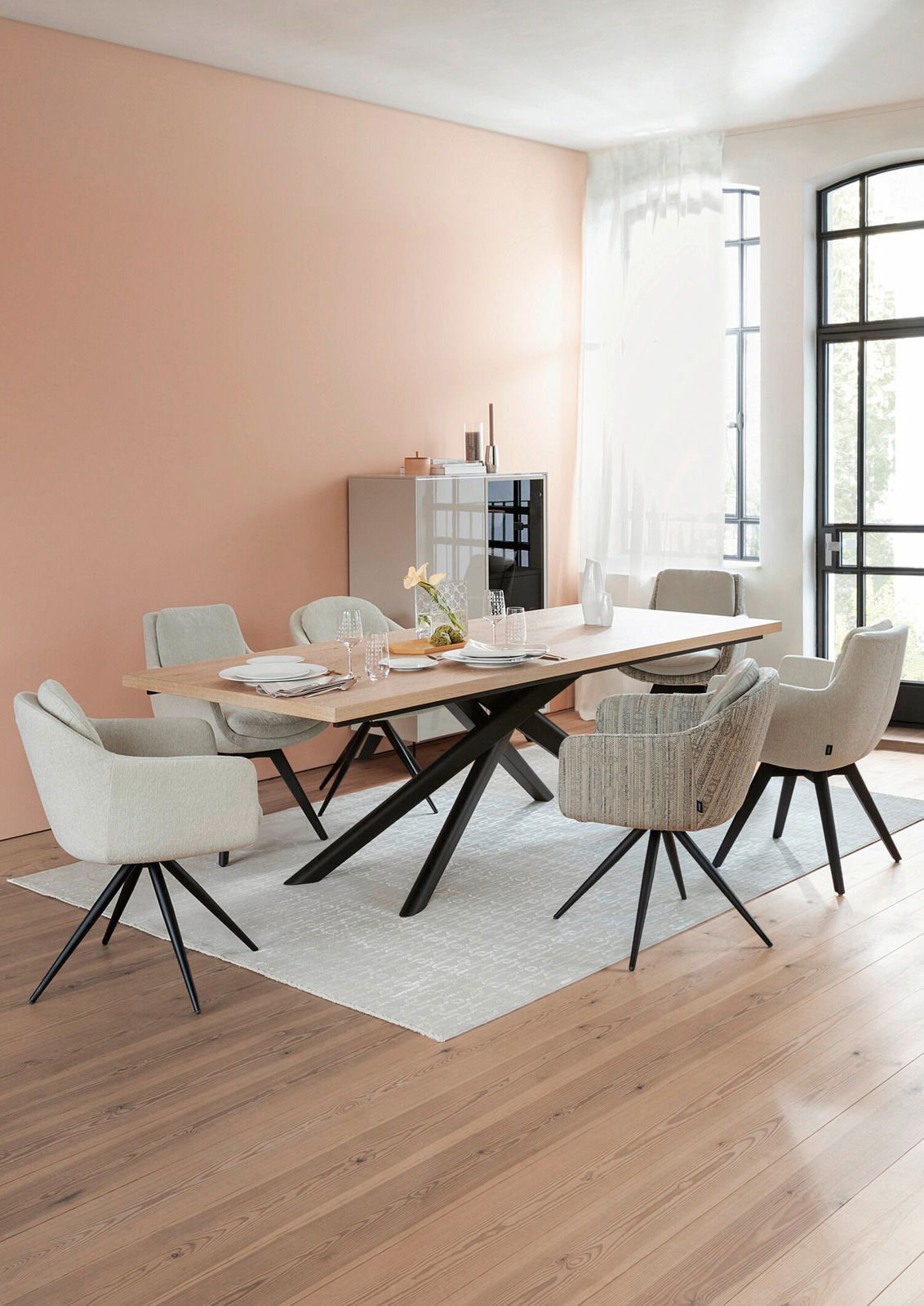 Moderner Esstisch mit Holzplatte und schwarzen Metallbeinen, umgeben von gepolsterten Stühlen in einem hellen Raum mit großen Fenstern, seitliche Perspektive.