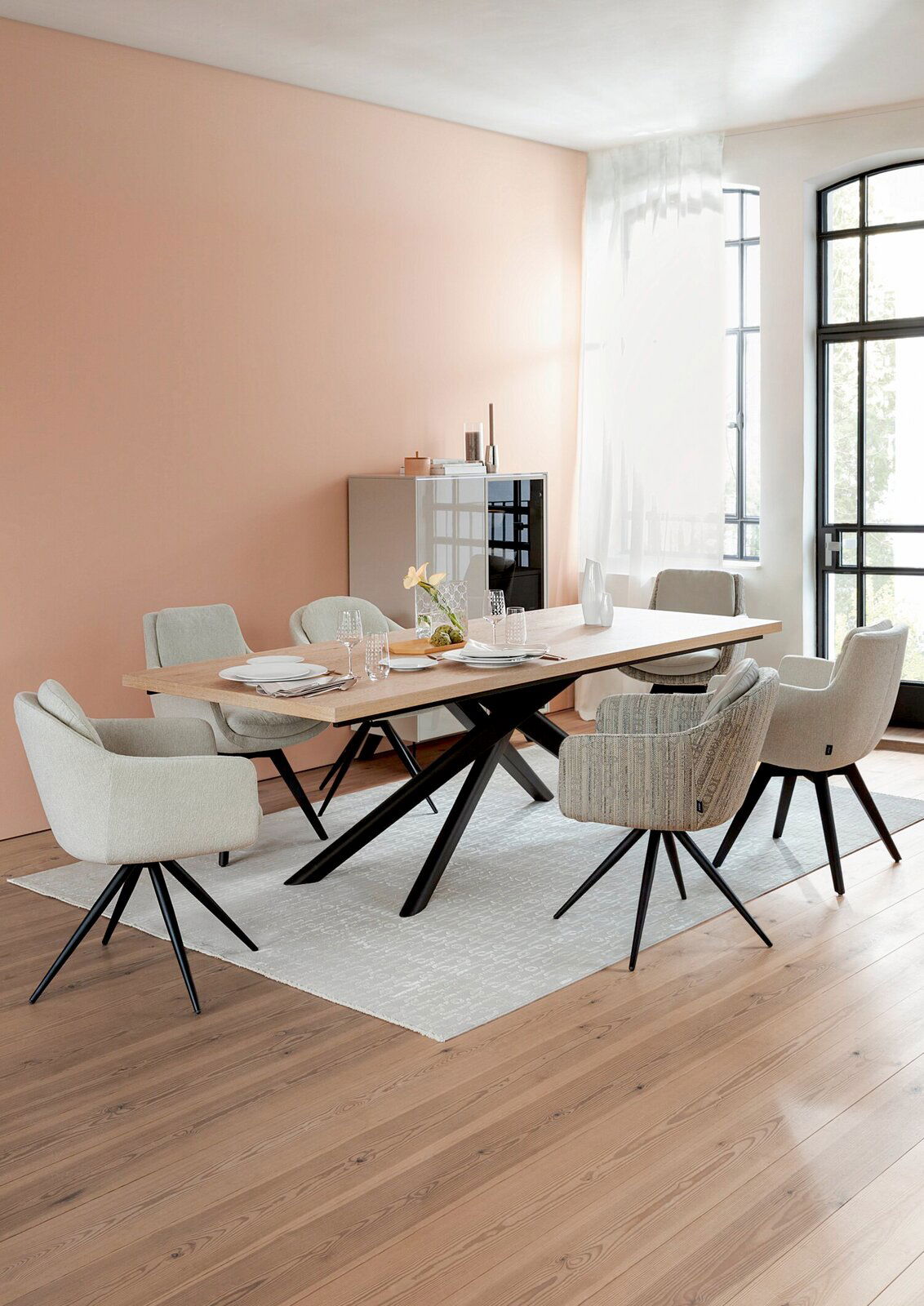 Moderner Esstisch mit Holzplatte und schwarzen Metallbeinen, umgeben von gepolsterten Stühlen in einem hellen Raum mit großen Fenstern, seitliche Perspektive.