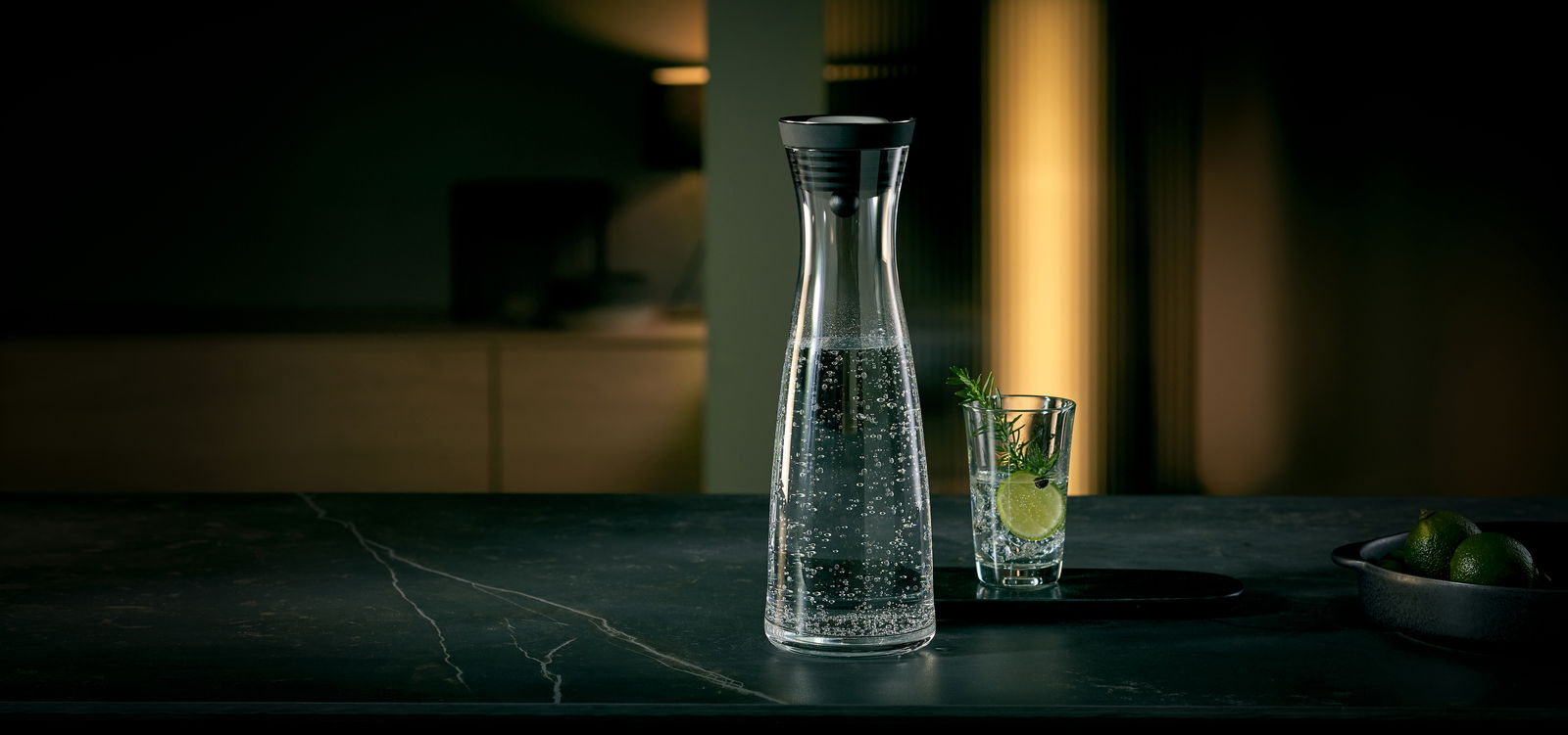 Elegante Wasserkaraffe BASIC 1,5L mit sprudelndem Wasser, seitliche Perspektive, neben einem Glas mit Limette und Rosmarin.