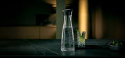 Elegante Wasserkaraffe BASIC 1,5L mit sprudelndem Wasser, seitliche Perspektive, neben einem Glas mit Limette und Rosmarin.