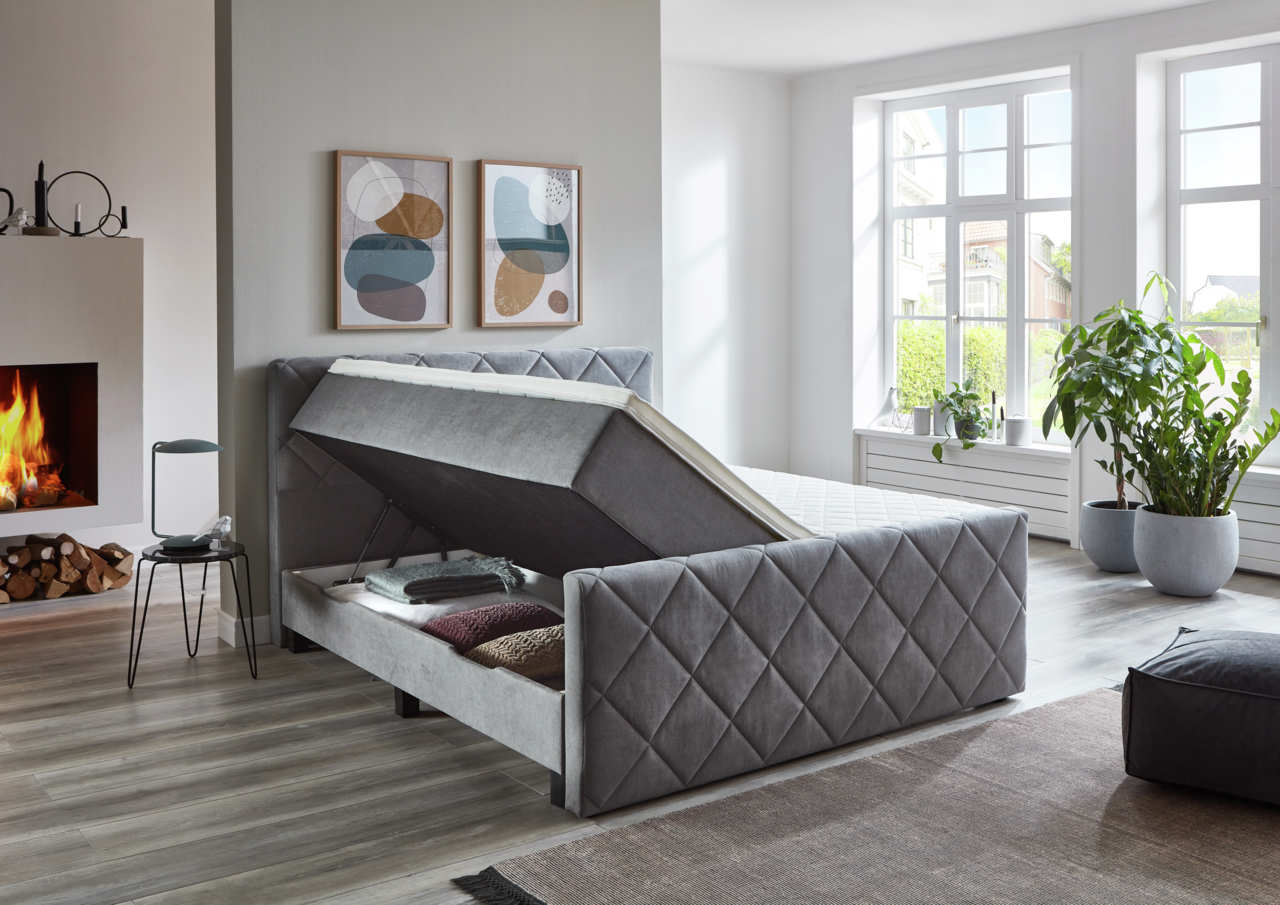 Graues Boxspringbett BX2200 CHESTER mit Stauraumfunktion, seitliche Perspektive, in modernem Schlafzimmer mit Kamin und großen Fenstern.