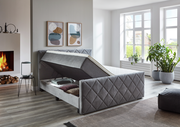 Graues Boxspringbett BX2200 CHESTER mit Stauraumfunktion, seitliche Perspektive, in modernem Schlafzimmer mit Kamin und großen Fenstern.