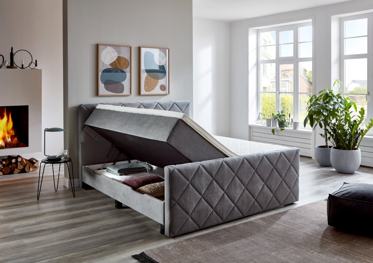 Graues Boxspringbett BX2200 CHESTER mit Stauraumfunktion, seitliche Perspektive, in modernem Schlafzimmer mit Kamin und großen Fenstern.