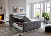 Graues Boxspringbett BX2200 CHESTER mit Stauraumfunktion, seitliche Perspektive, in modernem Schlafzimmer mit Kamin und großen Fenstern.