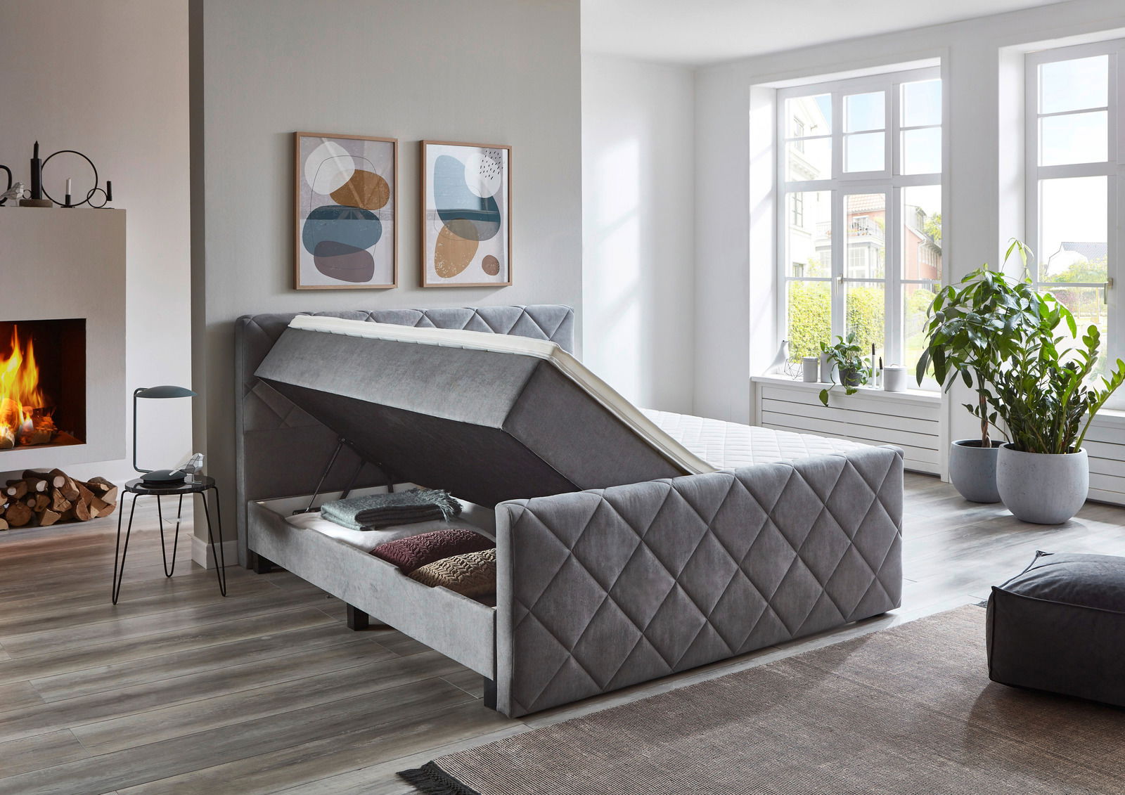 Graues Boxspringbett BX2200 CHESTER mit Stauraumfunktion, seitliche Perspektive, in modernem Schlafzimmer mit Kamin und großen Fenstern.