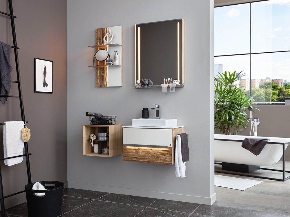 Das Bild zeigt ein modernes Badezimmer. Es gibt ein Waschbecken mit einem Holz-Unterschrank und einem großen Spiegel mit Beleuchtung. An der Wand hängen kleine Regale für Dekoration und Haushaltsgeräte. Im Hintergrund steht eine weiße Badewanne vor einem