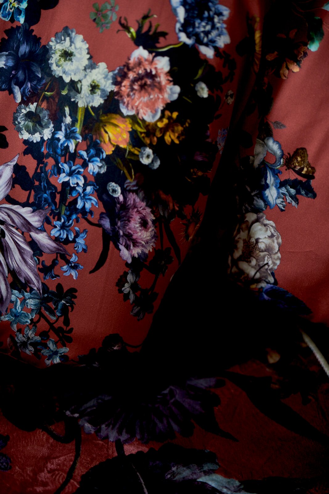 Nahaufnahme aus frontaler Perspektive auf einen drapierten Stoff mit opulentem Blumenmuster in Blau, Weiß, Rosa und Violett auf dunkelrotem Grund.