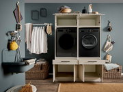 Frontale Perspektive: moderner Waschturm/Waschmaschinen-Schrank in Weiß mit zwei schwarzen Frontlader-Geräten (Waschmaschine und Trockner) in einem stilvollen Hauswirtschaftsraum, mit offenen Schranktüren unten und Ablagefläche oben.