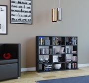 Frontale Innenraumansicht eines modernen Wohnzimmers: schwarzes Regal mit Büchern und Deko, darüber zwei zylindrische Pendelleuchten in Gold und Silber vor grauer Wand