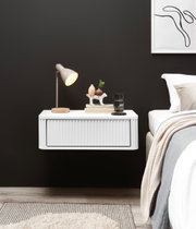 Zwevend wit nachtkastje met geribbeld front naast een bed, versierd met een tafellamp, boeken en een glas – vooraanzicht vanuit een licht zijdelings perspectief.
