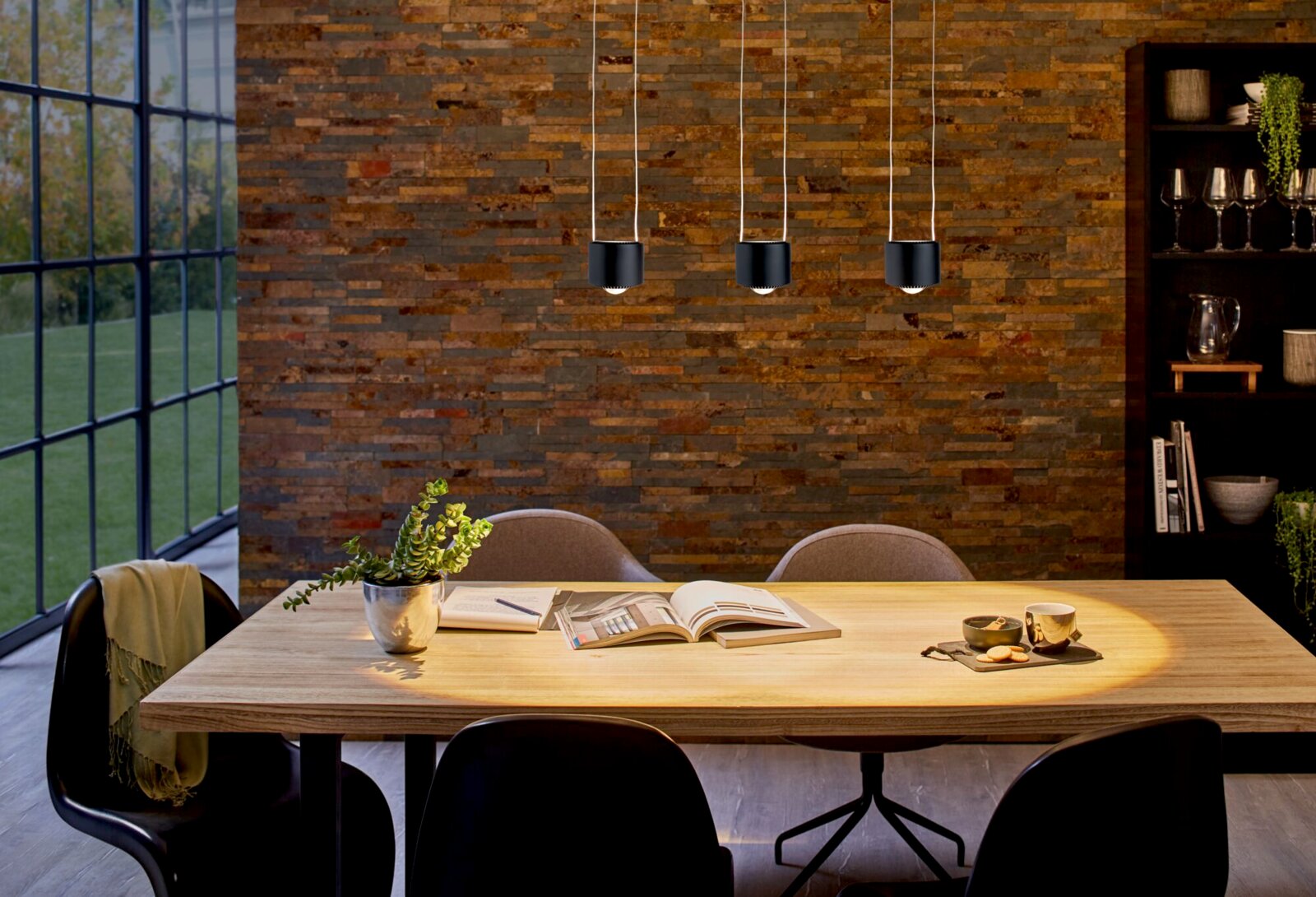 Drei moderne, schwarze Pendelleuchten über einem Esstisch in einem stilvoll eingerichteten Raum mit Backsteinwand. Perspektive: Frontalansicht auf die Leuchten und den Tisch.