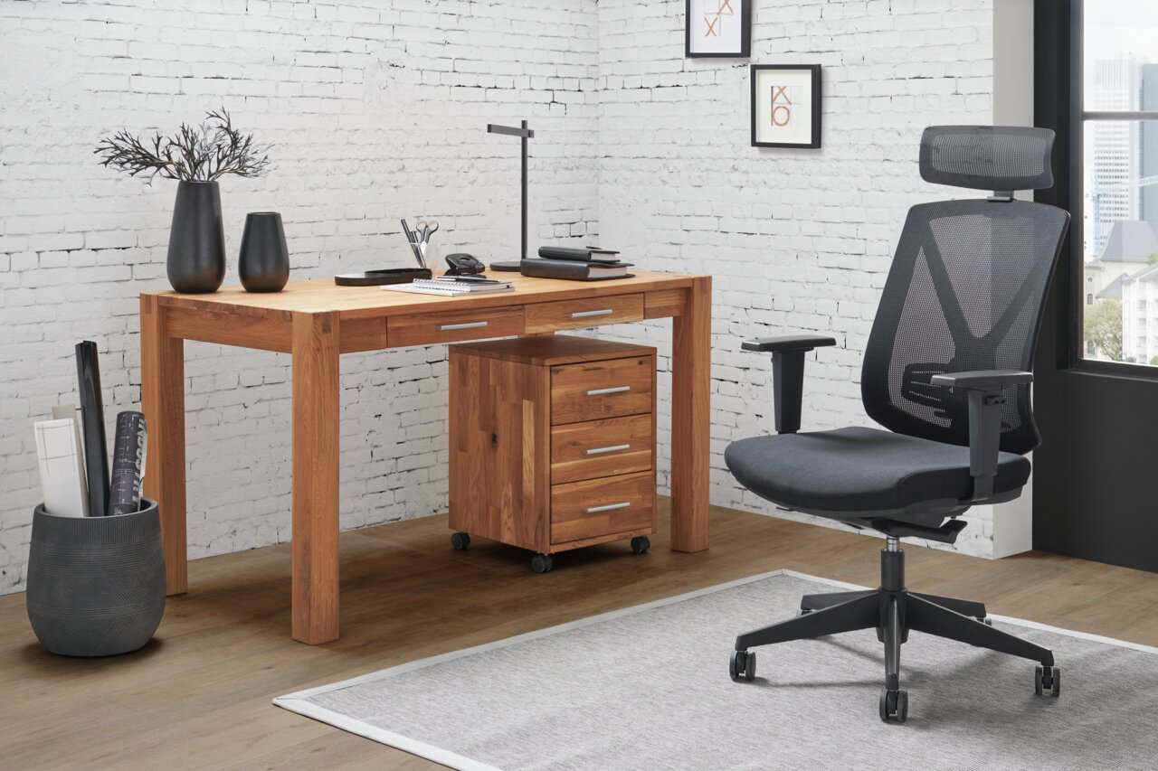 Moderner Schreibtisch aus Holz mit Schubladen, seitliche Perspektive, neben einem ergonomischen Bürostuhl in einem stilvollen Büro mit weißer Backsteinwand.