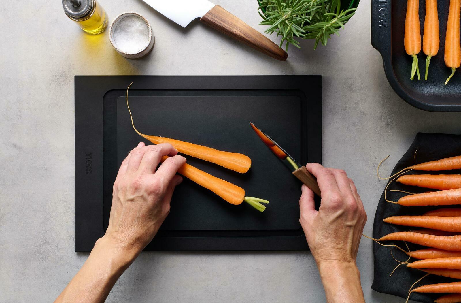 Schneidebrett aus schwarzem Material, auf dem eine Person Karotten mit einem Messer schneidet. Perspektive von oben.