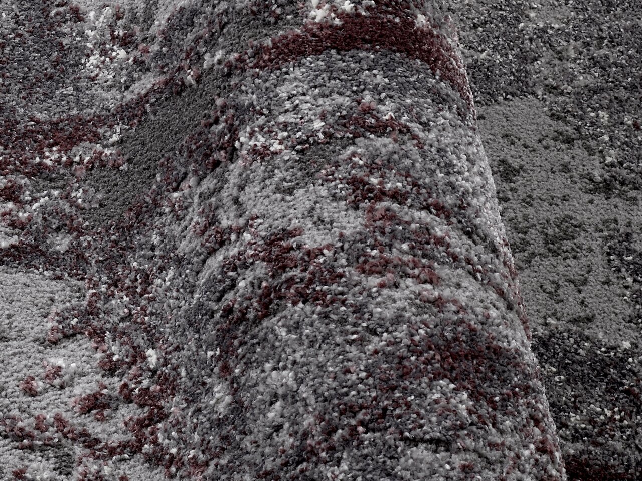 Nahaufnahme eines strukturierten Teppichs in Grautönen mit roten Akzenten, perspektivisch von der Seite aufgenommen.