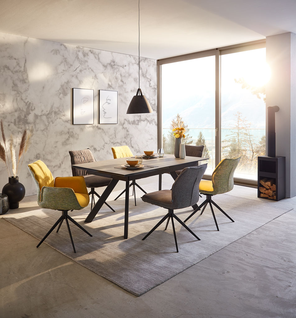 Novel Sessel HAPPY Esszimmer mit modernem Tisch und Stühlen in verschiedenen Farben, seitliche Perspektive mit Blick auf große Fenster und sonnige Aussicht.