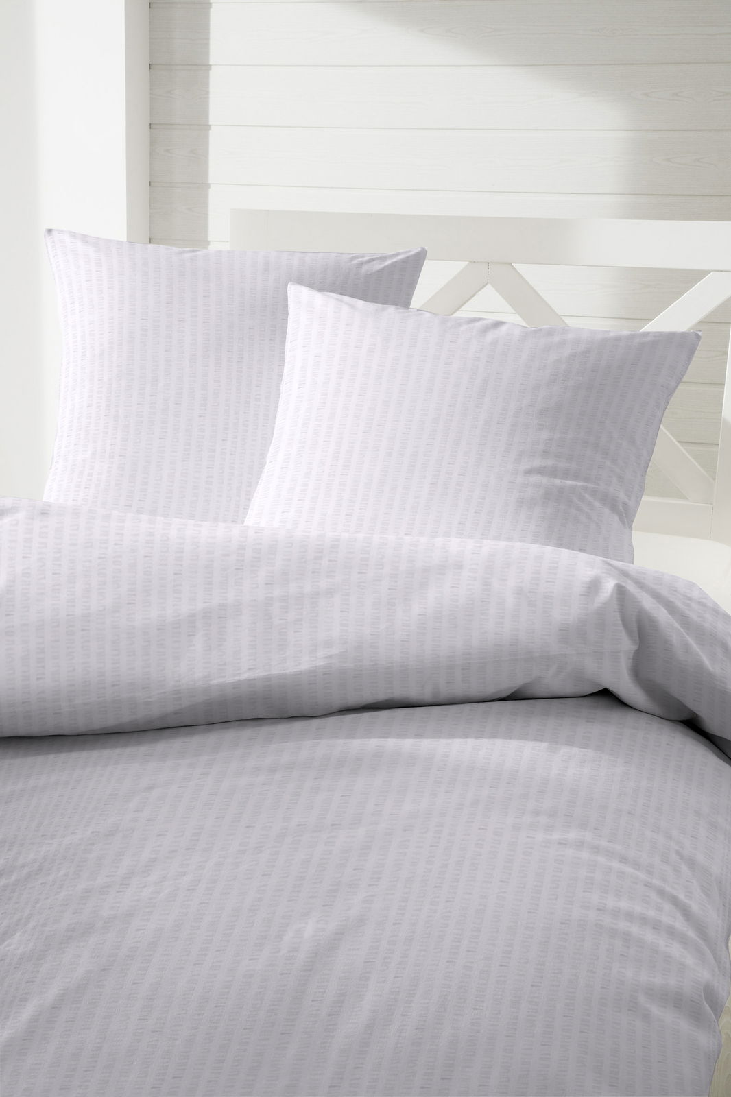 Weiße Bettwäschegarnitur mit dezentem Muster, bestehend aus einem Bettbezug und zwei Kissenbezügen, auf einem Bett mit weißem Kopfteil, aus der Perspektive von vorne fotografiert.