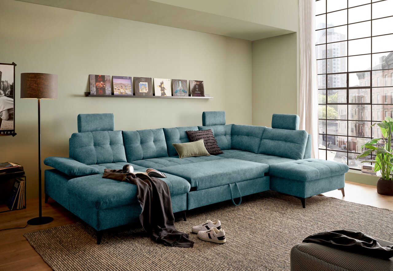 Großes, modernes U-förmiges Sofa in Türkis mit verstellbaren Kopfstützen, in einem stilvoll eingerichteten Wohnzimmer mit Blick auf eine Stadtlandschaft durch ein großes Fenster. Perspektive von vorne links.