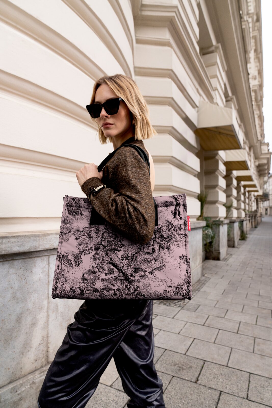 Frau mit Sonnenbrille trägt eine stilvolle, gemusterte Einkaufstasche in einer städtischen Umgebung. Die Tasche hat ein elegantes Design mit einem schwarz-weißen Muster. Perspektive von der Seite.