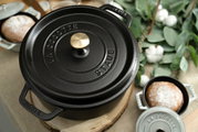 Schwarzer runder Bräter Cocotte 24cm von oben, mit goldfarbenem Knauf, umgeben von Brot und grünen Blättern.