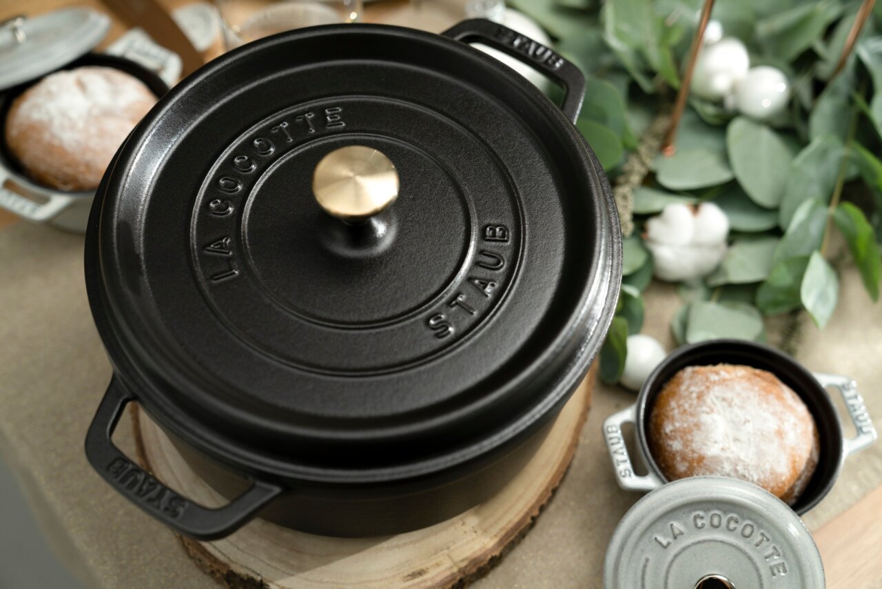 Schwarzer runder Bräter Cocotte 24cm von oben, mit goldfarbenem Knauf, umgeben von Brot und grünen Blättern.