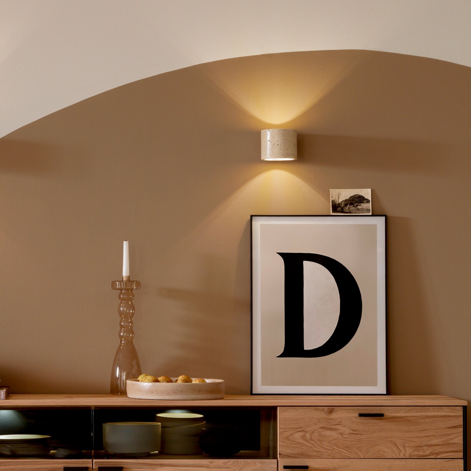 Beige zylindrische Wandleuchte mit gesprenkelter Oberfläche, die warmes Licht nach oben und unten abstrahlt, frontal in leichter Untersicht an einer sandfarbenen Wand über einem Sideboard inszeniert.