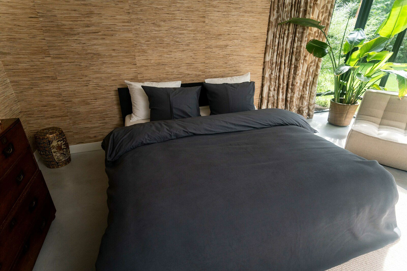 Kayori Bettwäsche ERIMO Blick auf ein stilvoll eingerichtetes Schlafzimmer mit einem großen Bett, das mit dunkelgrauer Bettwäsche in der Größe 155x220cm bezogen ist. Im Hintergrund sind eine beige Wand, Vorhänge und eine große grüne Pflanze zu sehen.