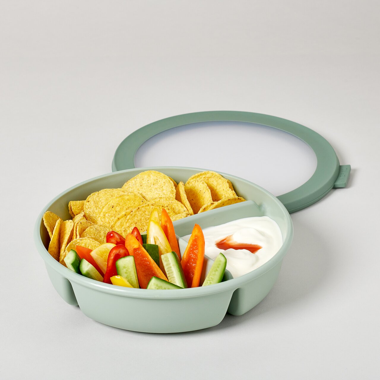 Grüne runde Bento-Schale Cirqula mit Deckel, gefüllt mit Chips, geschnittenem Gemüse und Dip, aus der Vogelperspektive.