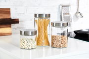 Drei runde Vorratsdosen aus Glas mit Edelstahl-Deckeln, gefüllt mit weißen Bohnen, Spaghetti und Kichererbsen, auf einer Küchenarbeitsplatte in seitlicher Perspektive.