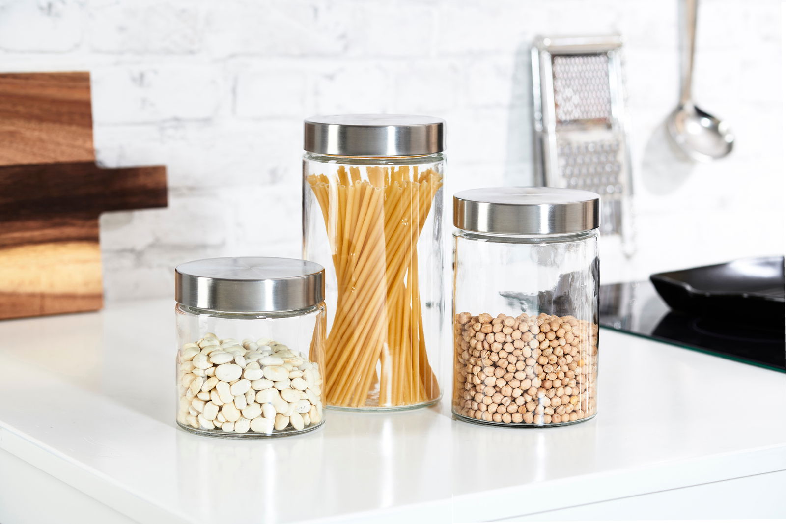 Drei runde Vorratsdosen aus Glas mit Edelstahl-Deckeln, gefüllt mit weißen Bohnen, Spaghetti und Kichererbsen, auf einer Küchenarbeitsplatte in seitlicher Perspektive.