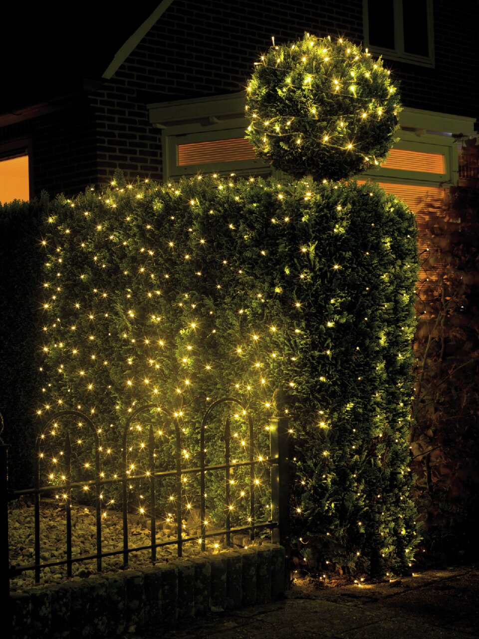 Een LED-lichtslinger verlicht ’s nachts een haag en een ronde struik in een tuin, gezien vanuit de zijkant.