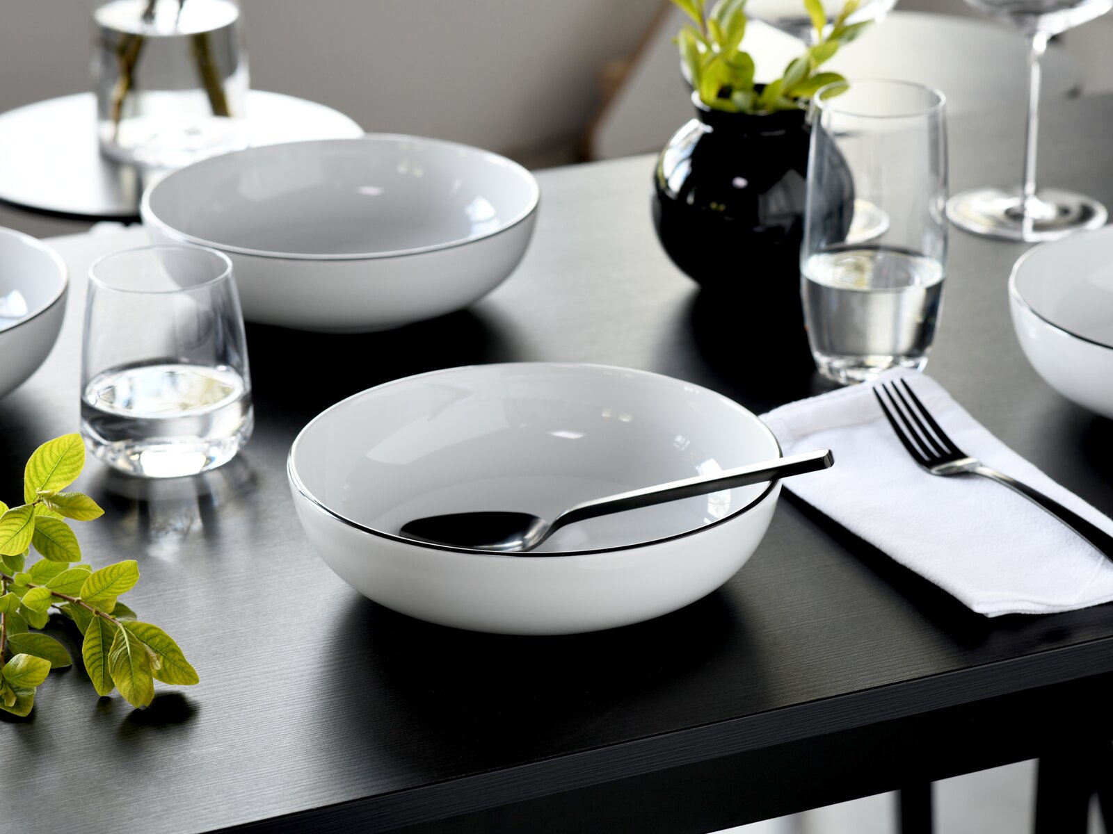 Modern gedeckter Esstisch mit weißen Porzellanschalen mit feinem dunklem Rand, Wassergläsern, Besteck und Serviette, fotografiert aus leicht erhöhter schräger Perspektive von vorne.