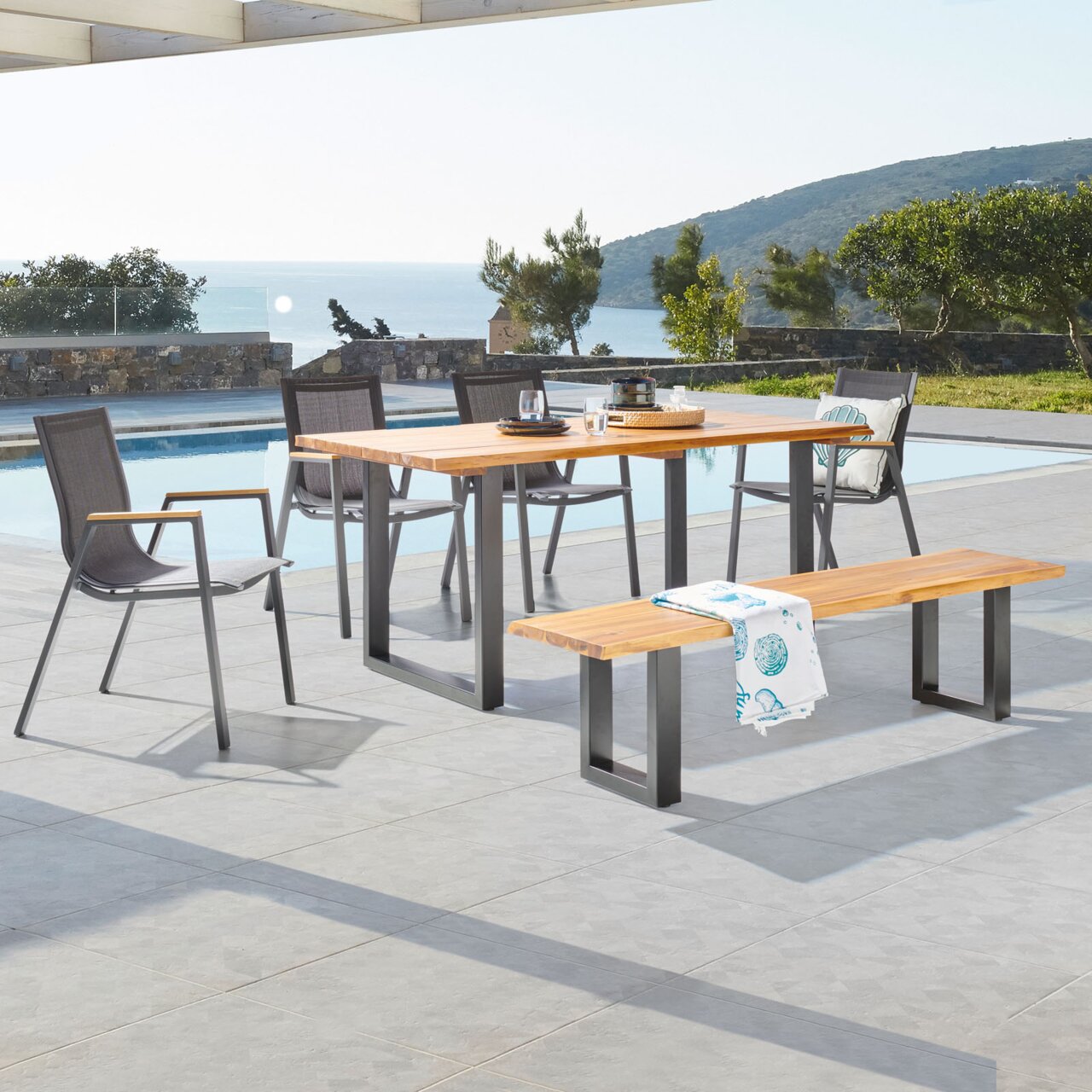 Ambia Garden Stapelsessel HONOLULU Outdoor-Esstisch-Set mit rechteckigem Holztisch auf schwarzen Metallkufen, vier grauen Gartenstühlen und passender Holzbank auf Poolterrasse mit Meerblick, perspektivisch in leichter Seitenansicht aufgenommen