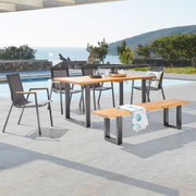 Outdoor-Esstisch-Set mit rechteckigem Holztisch auf schwarzen Metallkufen, vier grauen Gartenstühlen und passender Holzbank auf Poolterrasse mit Meerblick, perspektivisch in leichter Seitenansicht aufgenommen