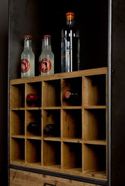 Nahaufnahme eines Weinregals aus Holz mit mehreren Fächern, in denen Weinflaschen horizontal gelagert sind. Oben stehen zwei Flaschen Ginger Beer und eine Flasche Gin. Perspektive von schräg vorne.