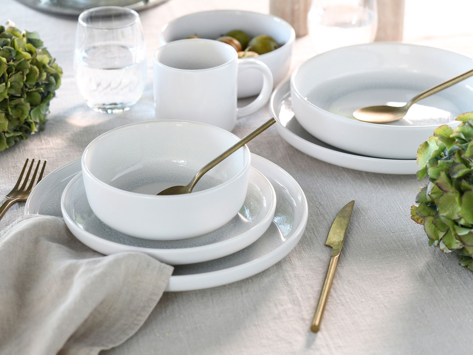 Elegantes weißes Kombiservice-Set mit Tellern, Schalen und Tassen, arrangiert auf einem gedeckten Tisch mit goldfarbenem Besteck und grünen Hortensien, aus der Vogelperspektive fotografiert.