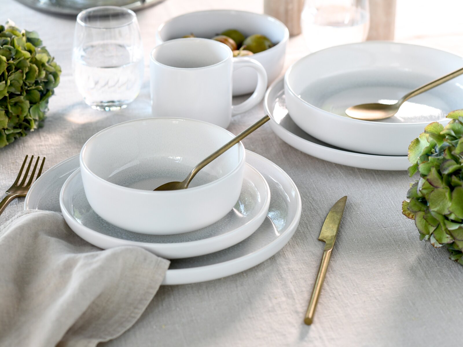 Elegantes weißes Kombiservice-Set mit Tellern, Schalen und Tassen, arrangiert auf einem gedeckten Tisch mit goldfarbenem Besteck und grünen Hortensien, aus der Vogelperspektive fotografiert.