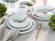 Elegantes weißes Kombiservice-Set mit Tellern, Schalen und Tassen, arrangiert auf einem gedeckten Tisch mit goldfarbenem Besteck und grünen Hortensien, aus der Vogelperspektive fotografiert.
