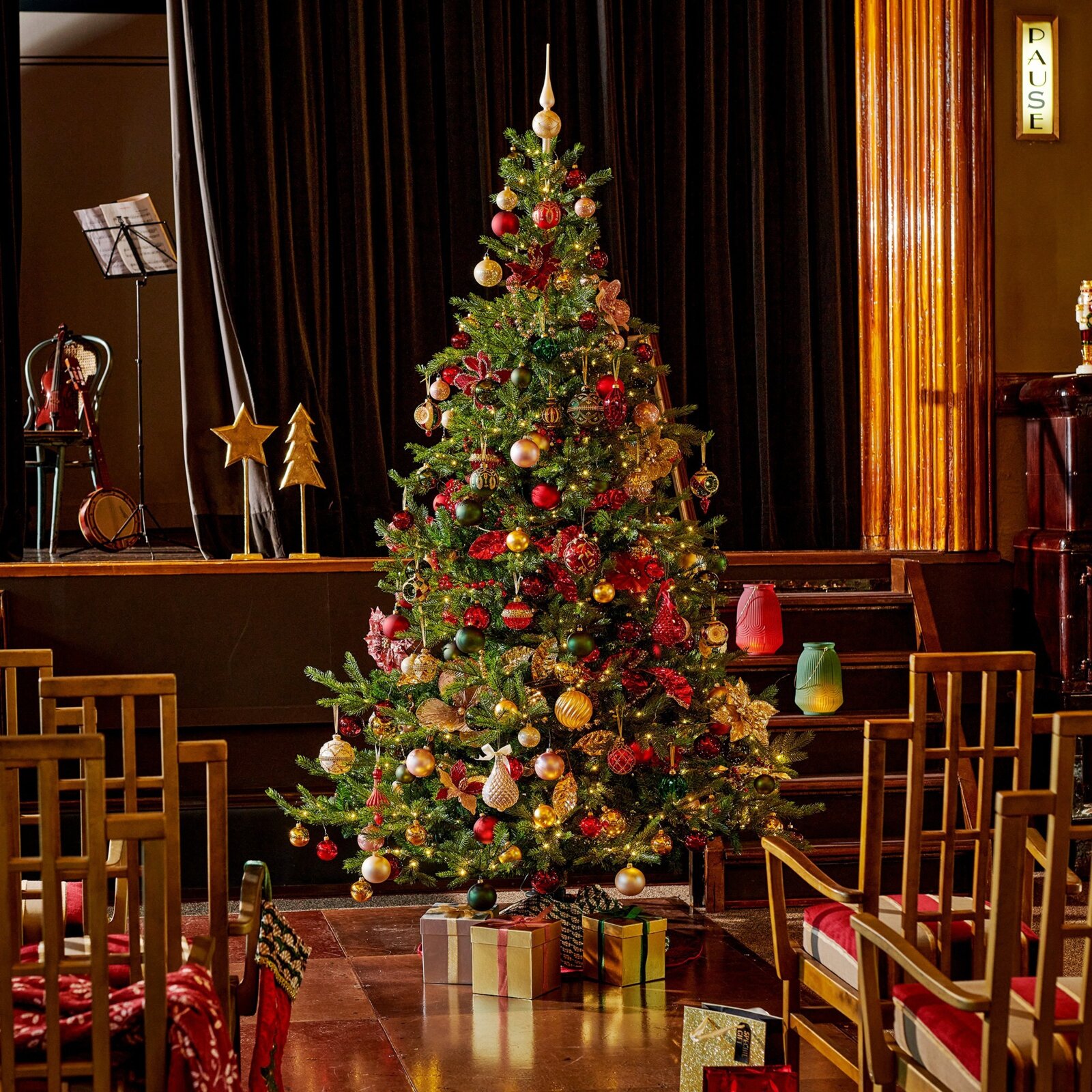 Frontaal totaalbeeld van een rijk versierde kerstboom met warme lichtjes, rode en gouden ornamenten en cadeaus in een sfeervolle theaterzaal.