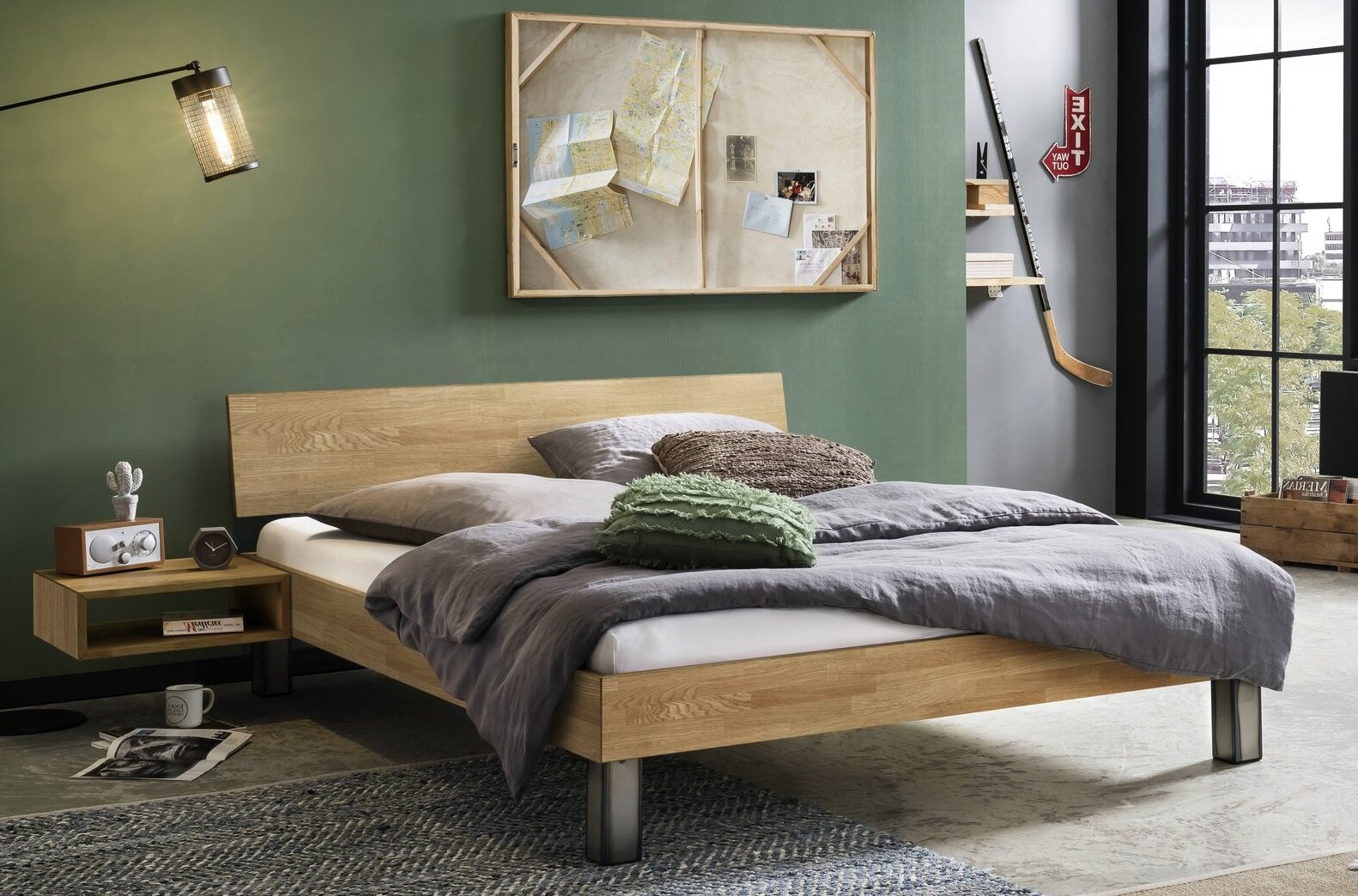 Modernes Holzbett mit schlichtem Kopfteil und Metallfüßen, grau bezogen und mit Kissen dekoriert, im Schlafzimmer-Setting Perspektive: frontale Seitenansicht leicht von rechts.