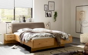 Modernes Doppelbett mit massivem Holzrahmen und braun gepolstertem Kopfteil in hellem Schlafzimmer, aufgenommen in schräger Frontansicht von links