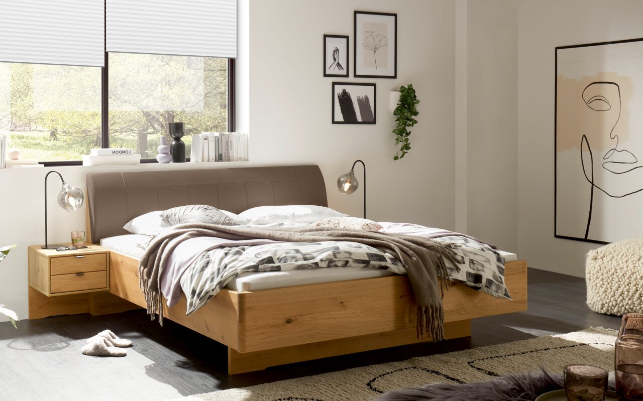Linea Natura Comfortabel bed CARDIFF Modern tweepersoonsbed met een massief houten frame en een bruin gestoffeerd hoofdeinde in een lichte slaapkamer, gefotografeerd vanuit een schuin vooraanzicht vanaf de linkerkant