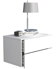 Weißer Nachttisch mit zwei Schubladen, obere Schublade leicht geöffnet; darauf silberne Tischlampe, Bücherstapel und kleiner Wecker, perspektivisch in schräger Vorderansicht von links.
